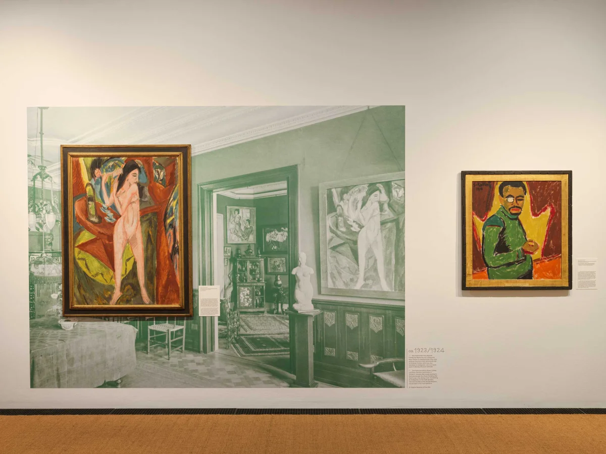Nude painting in front of a large green-tinted interior photograph, beside a portrait painting on white.