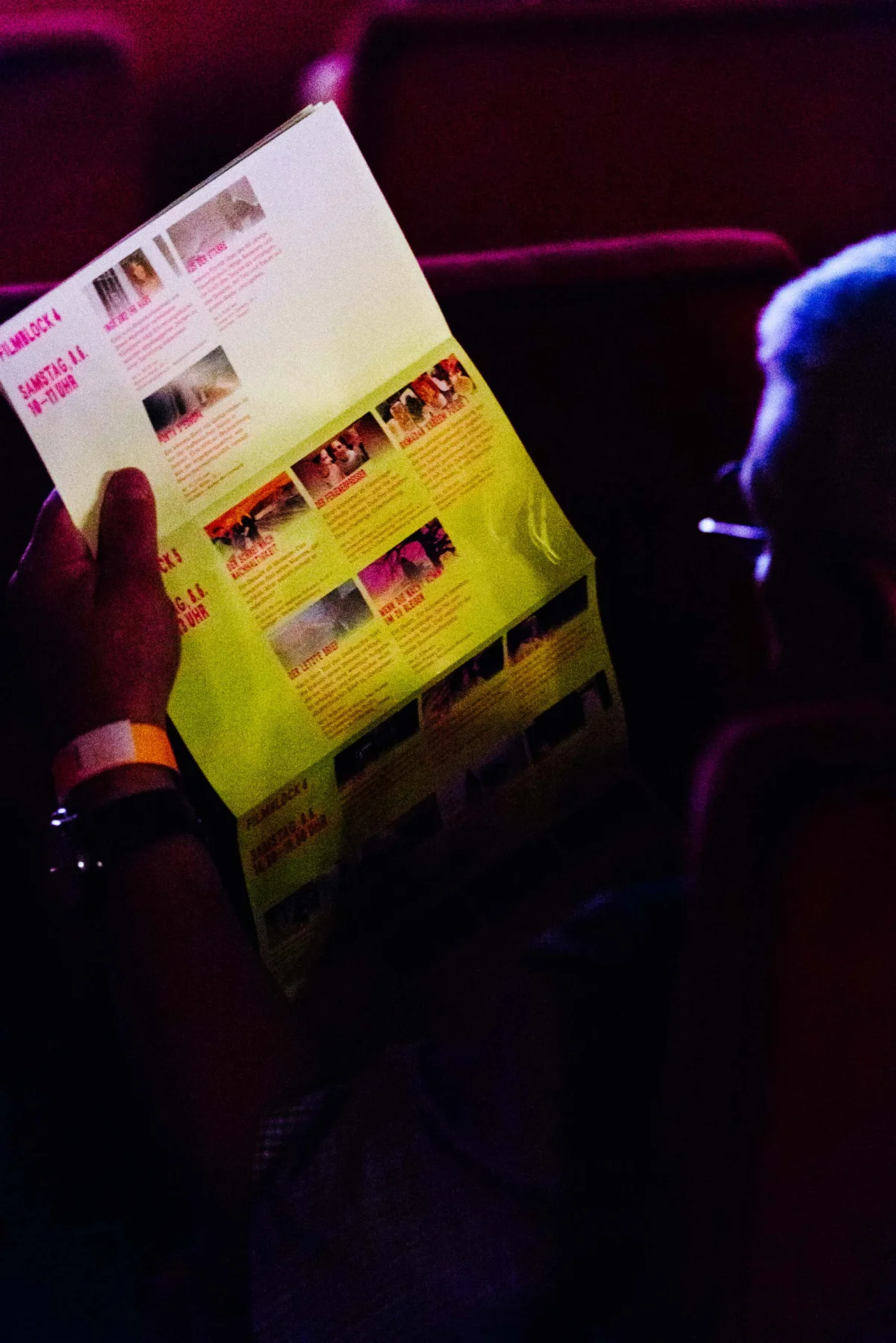 A person in a dark cinema seat holds an unfolded programme with film stills and a timetable.