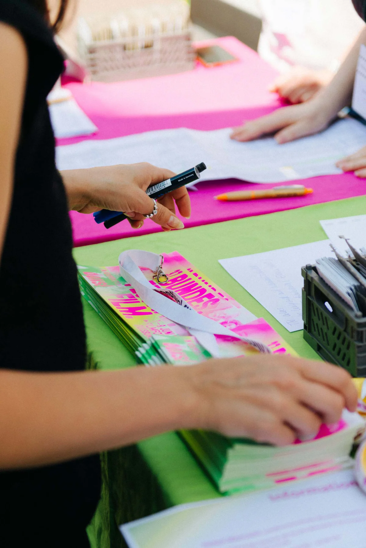 Hands and pens above printed festival programmes on a pink and green tabletop.