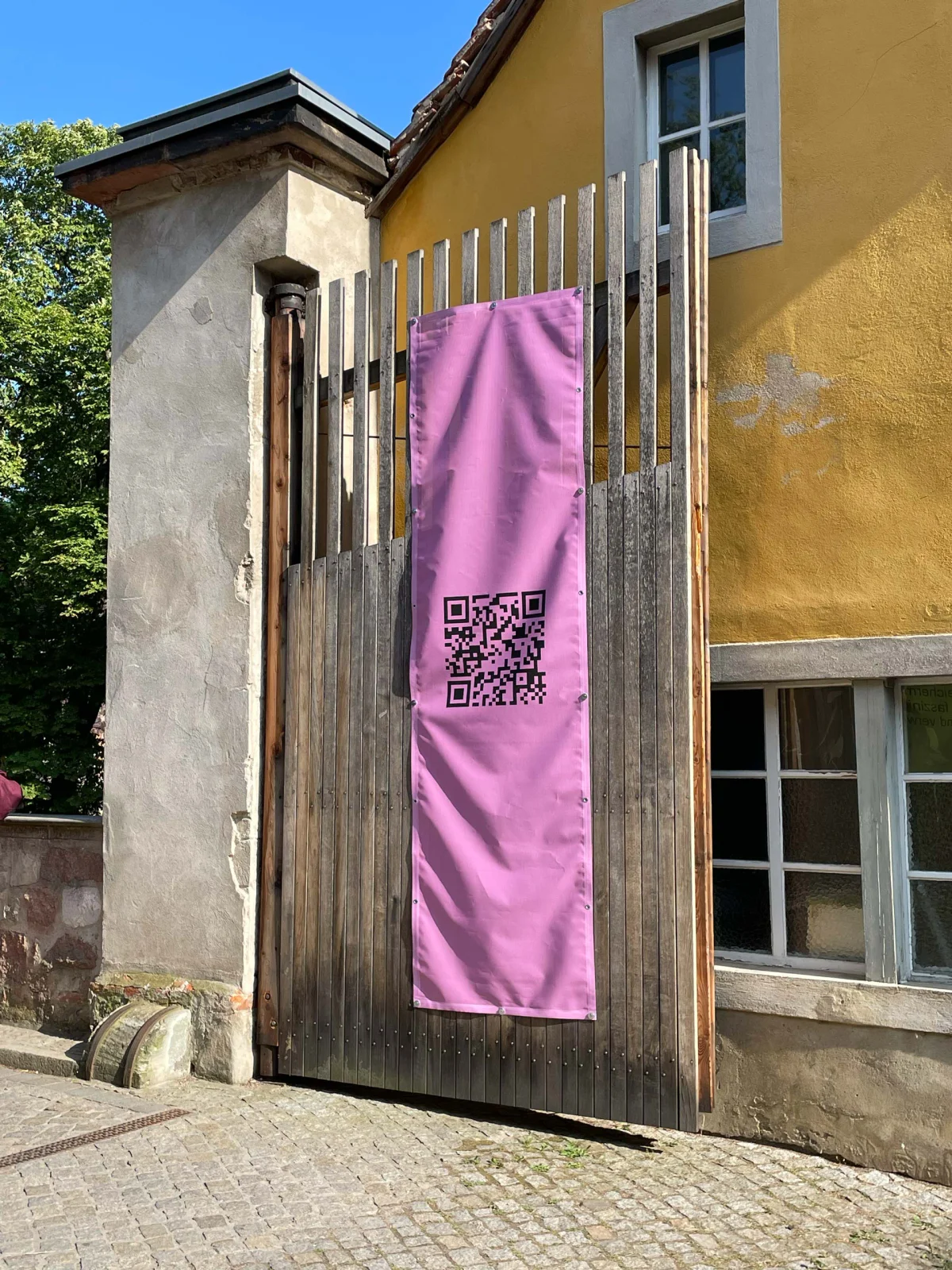 Pink banner with a QR code hanging on a wooden fence outside a yellow building.