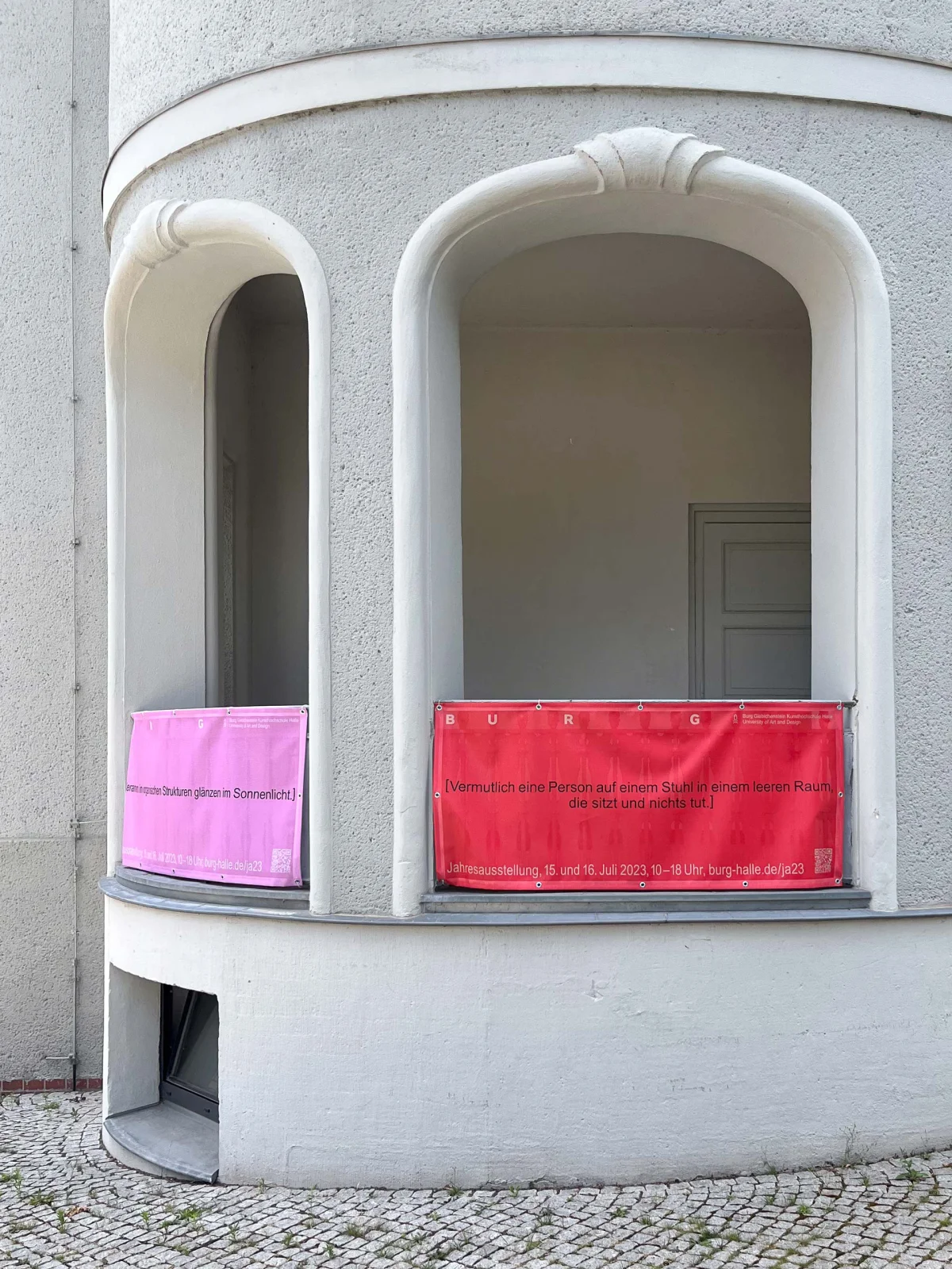 Pink and red BURG annual exhibition text banners displayed in two windows of a pale facade.
