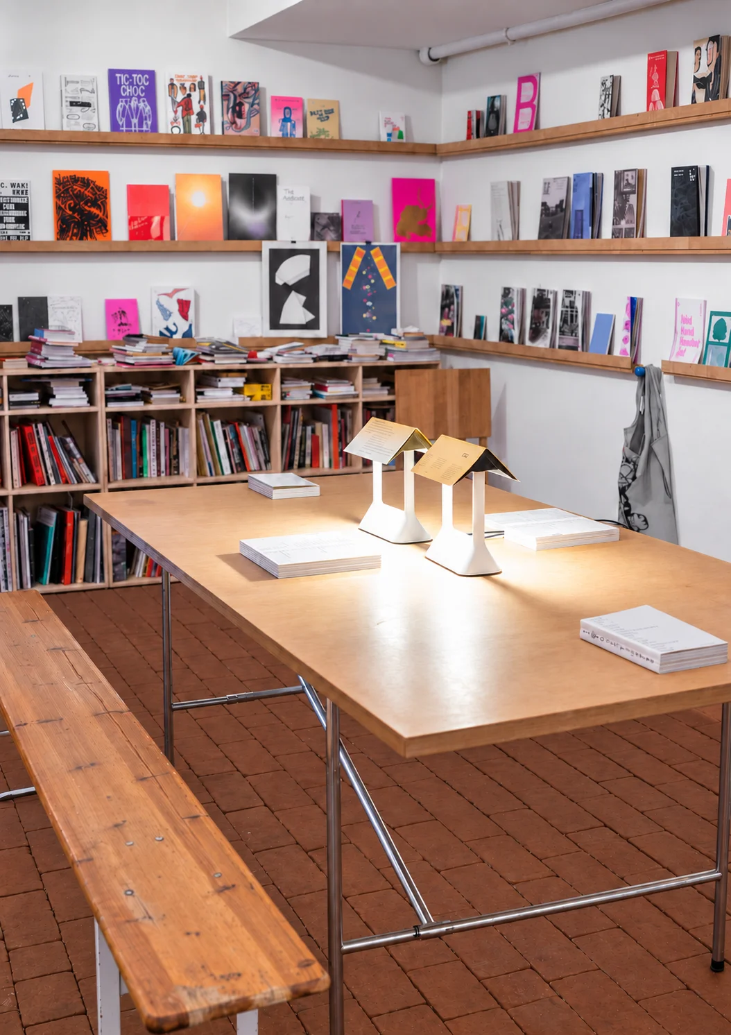 Large worktable with a wooden bench, two lit table lamps and books in front of shelves filled with publications.