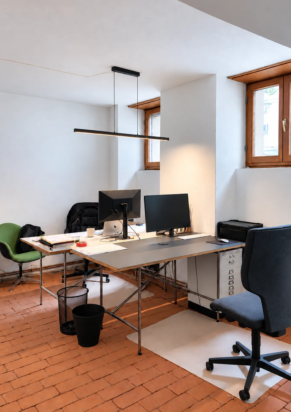 Studio workstations with two monitors, office chairs and a pendant light against white walls with two windows.