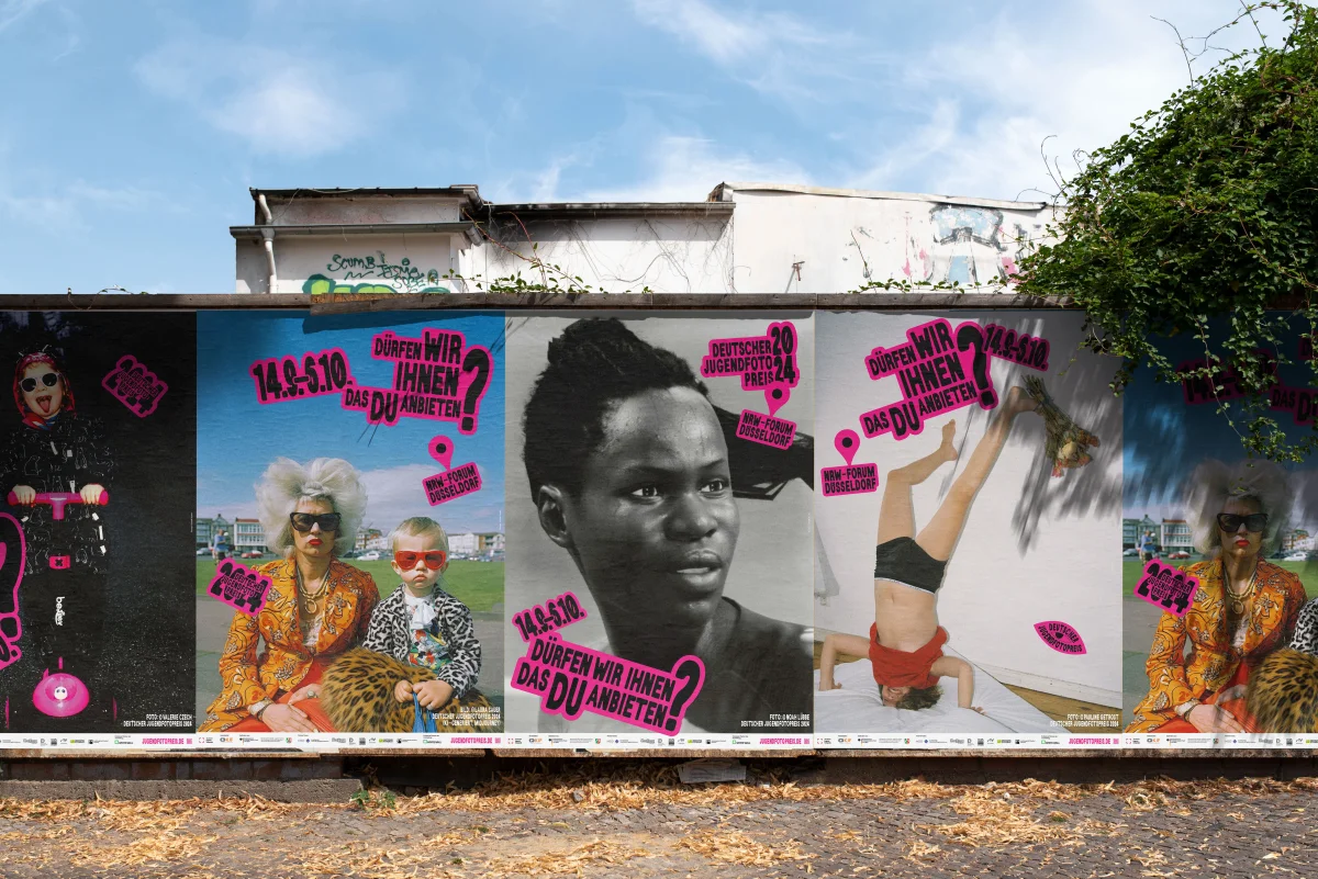 Four adjacent Youth Photography Award posters with different photographs and pink stickers on a wall above a pavement.