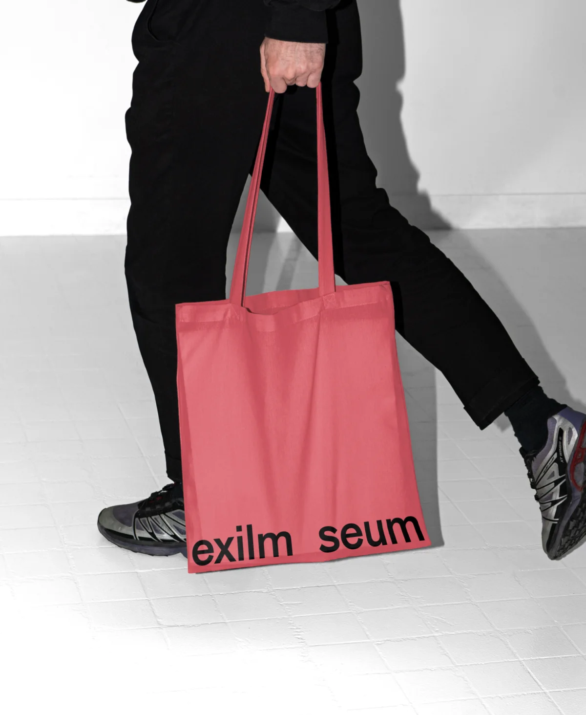 A walking person carries a coral-red tote bag with a black Exilmuseum wordmark along the bottom edge.