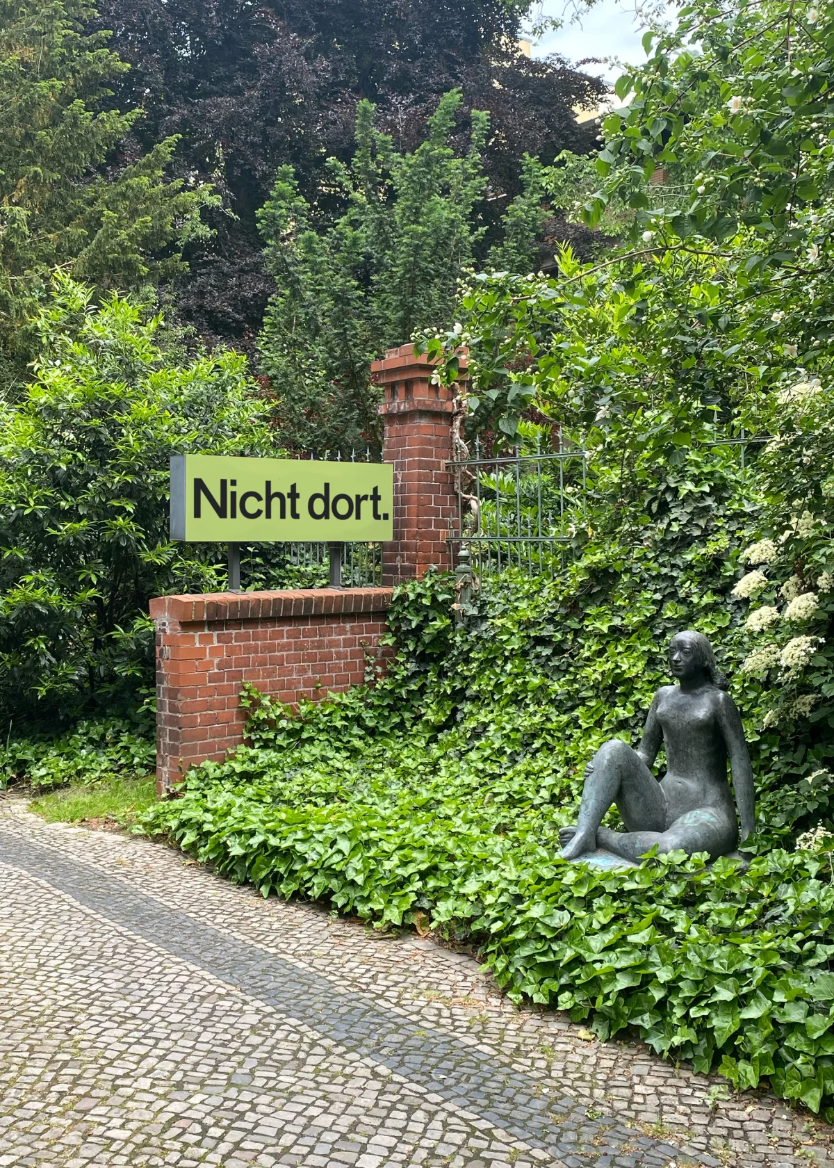 Yellow-green sign reading “Nicht dort.” on a brick wall in a planted garden beside a seated bronze sculpture.