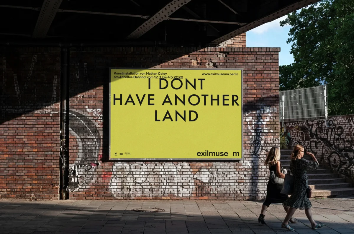 Yellow Exilmuseum poster reading “I DON'T HAVE ANOTHER LAND” on a graffiti-covered brick wall beneath a bridge.