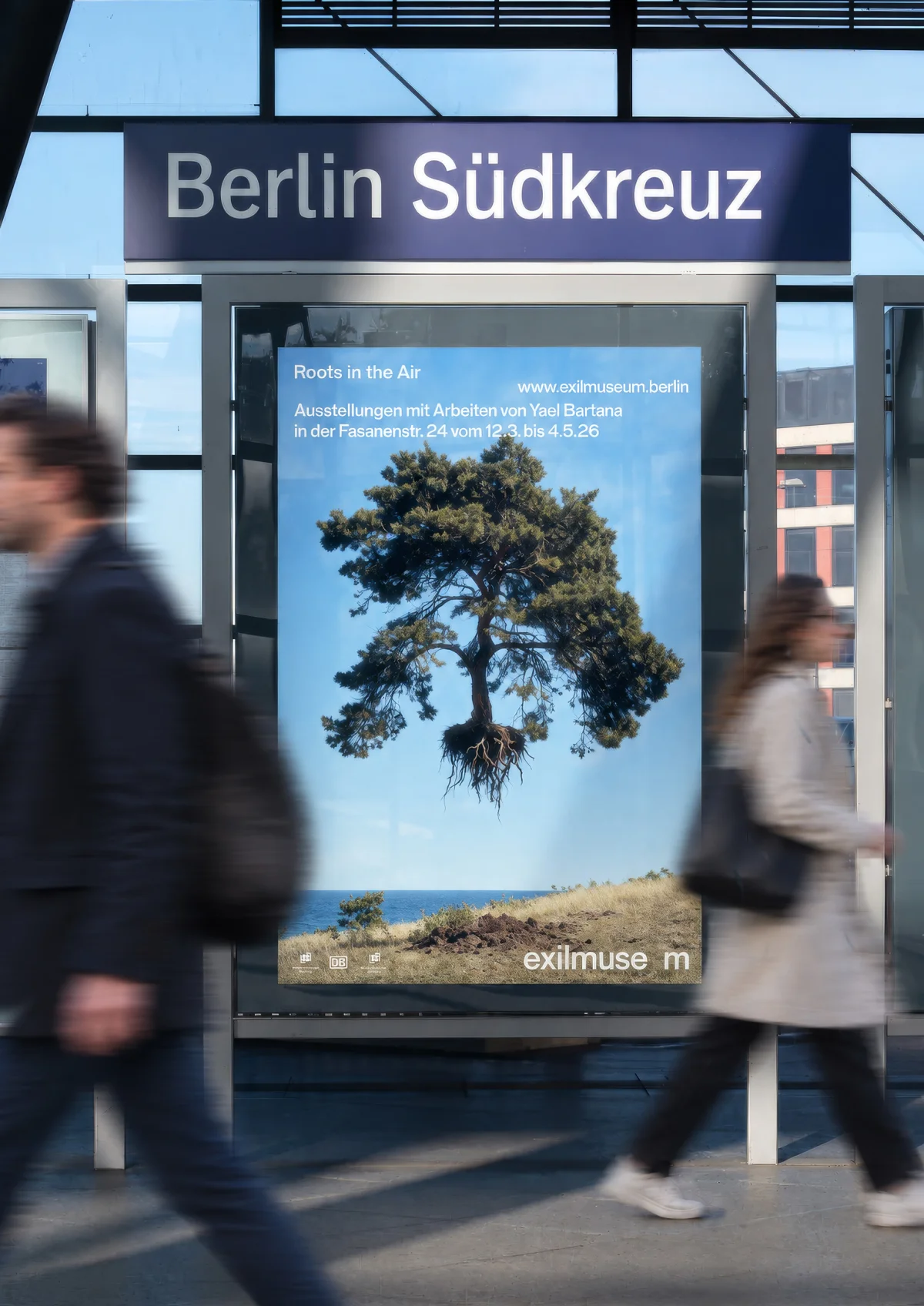 People walk past an advertising display at Berlin Südkreuz station, where a blue Exilmuseum poster shows a floating tree.