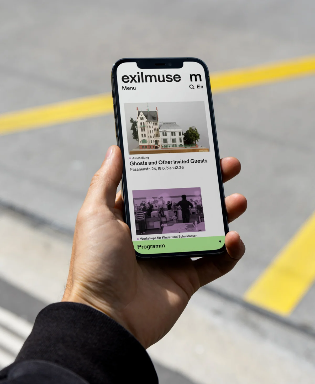 A hand holds a smartphone displaying the Exilmuseum website, an architectural model, an exhibition preview and a green programme bar.
