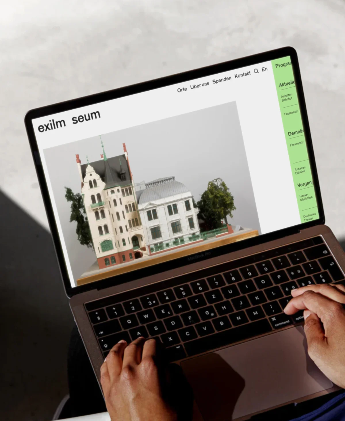 Hands operate a laptop displaying the Exilmuseum website, with an image of an architectural model and a green programme column.