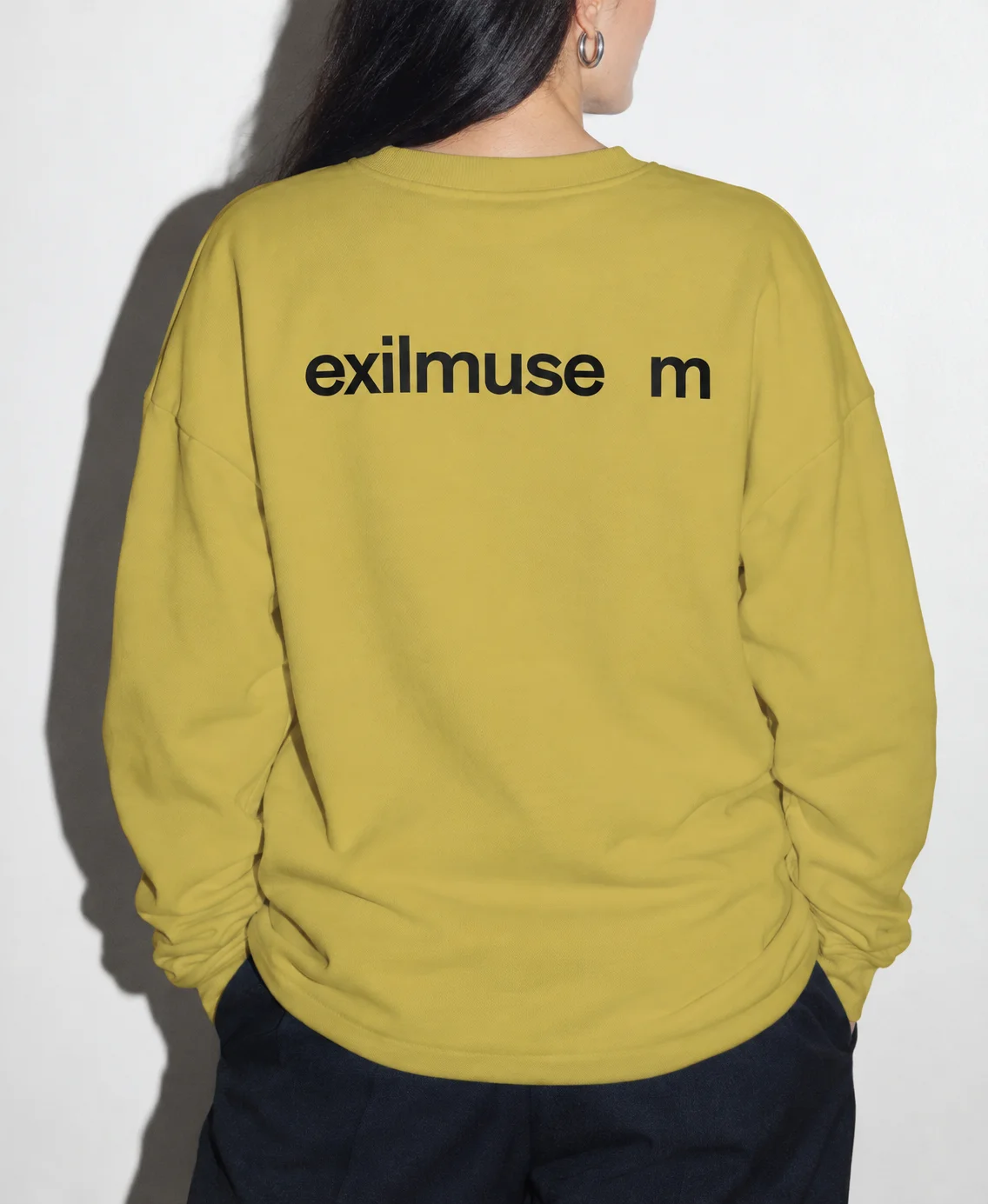 Back view of a person wearing a yellow-green sweatshirt with a black Exilmuseum wordmark and a gap before the m.