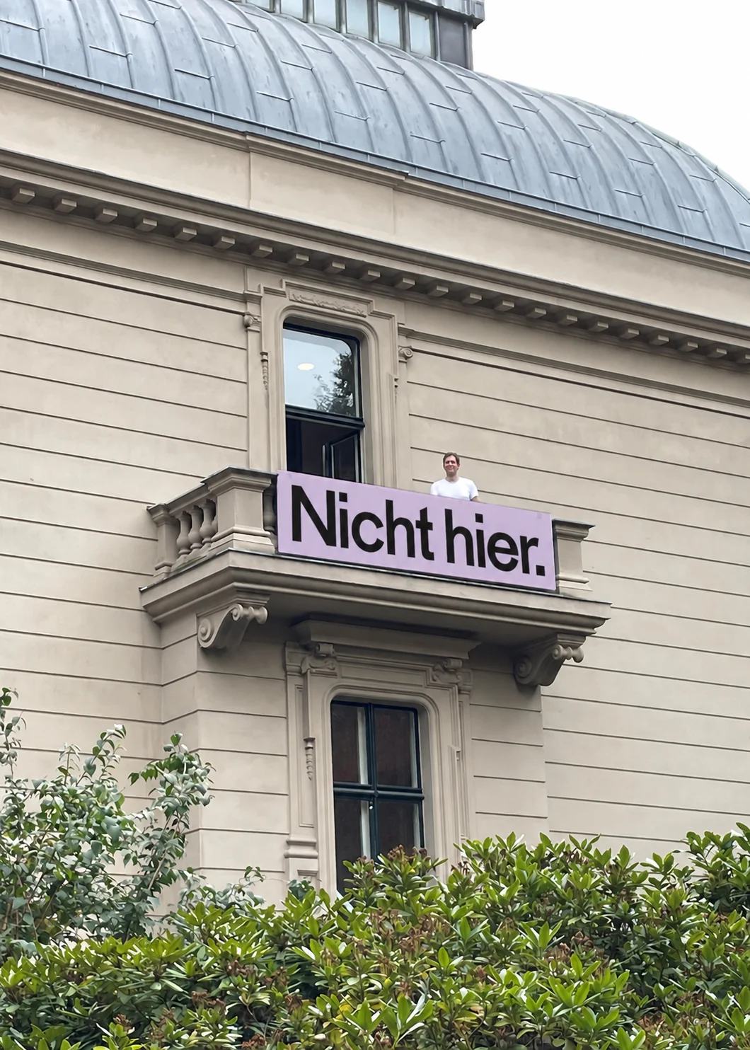 Pink banner reading “Nicht hier.” on the balcony of a pale historic building, with a person standing behind it.