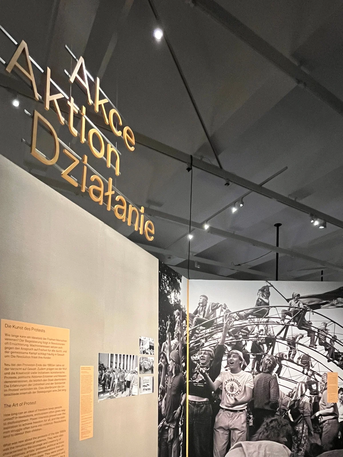 Gelber, dreisprachiger Titel „Aktion“ über einer Ausstellungswand mit Texttafeln und einer großformatigen Schwarz-Weiß-Fotografie.
