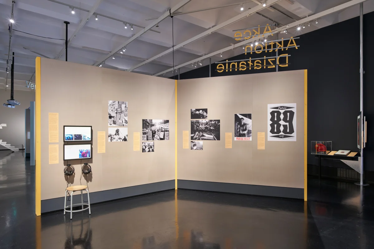 Ausstellungsbereich „Aktion“ mit gelben Wandkanten und Texttafeln, Schwarz-Weiß-Fotos, zwei Bildschirmen, Kopfhörern und Sitzhocker.