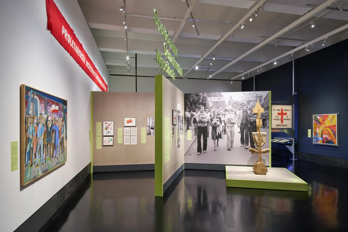 Weiter Blick in den Bereich „Gemeinschaft“ mit Gemälden, rotem Banner, großem Schwarz-Weiß-Gruppenfoto und einer Skulptur.