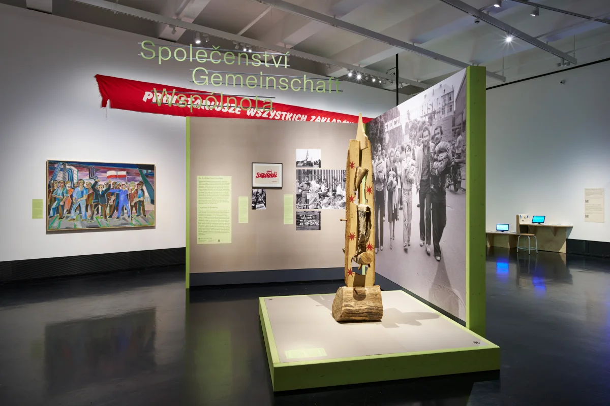 Ausstellungsbereich „Gemeinschaft“ mit grünen Wandkanten, großem Gruppenfoto und einer geschnitzten Skulptur auf einem hellgrünen Sockel.