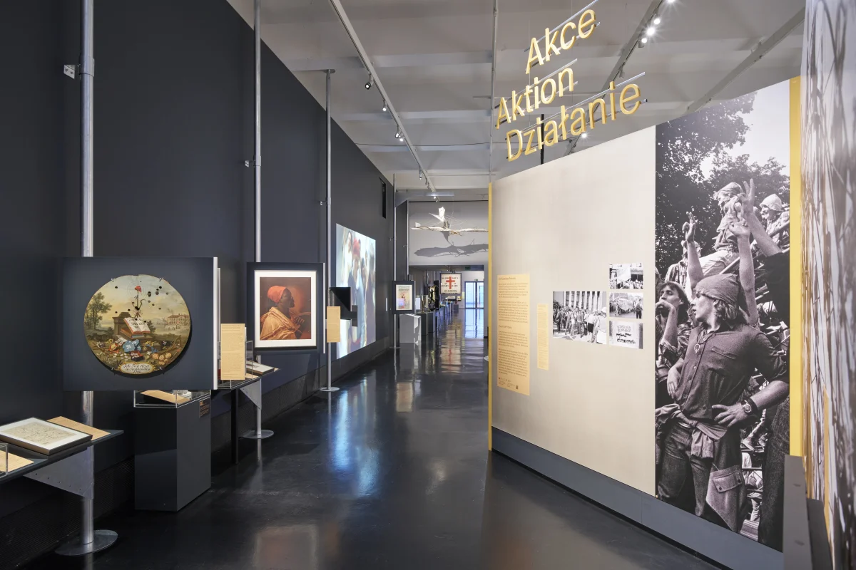 Blick durch den Ausstellungsbereich „Aktion“ mit beigefarbenen Wänden und einer großen Schwarz-Weiß-Aufnahme einer Menschenmenge.