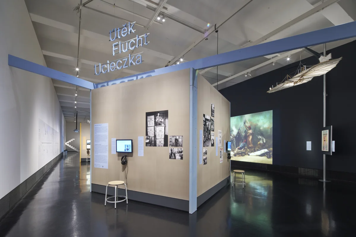 Ausstellungsbereich „Flucht“ mit blau umrahmten Stellwänden, Foto- und Texttafeln, einer Hörstation und einem über dem Raum hängenden Objekt.