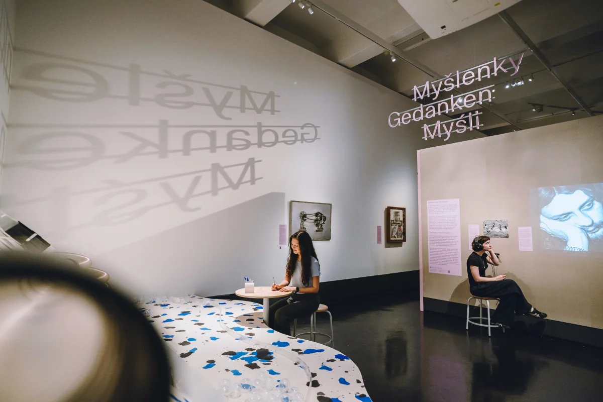 Zwei Personen sitzen im Ausstellungsbereich „Gedanken“, eine an einem gemusterten Tisch, eine mit Kopfhörern vor einer Bildprojektion.