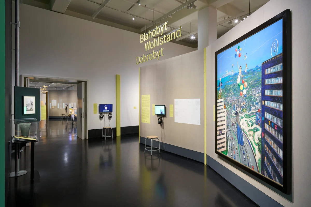 Ausstellungsbereich „Wohlstand“ mit gelben Wandkanten, Bildschirmen und Kopfhörern sowie einem großen farbigen Gemälde.