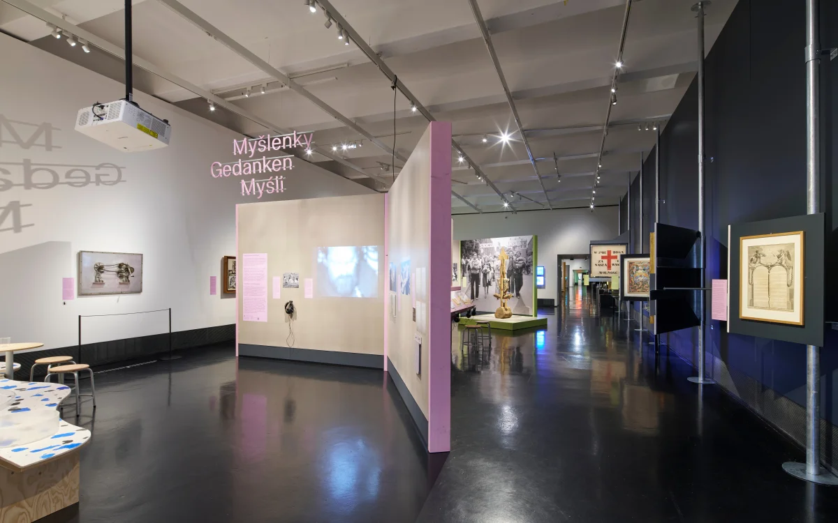 Ausstellungsraum mit frei stehenden beigefarbenen Stellwänden, rosa Informationstafeln, Bildern und dem mehrsprachigen hängenden Titel „Gedanken“.