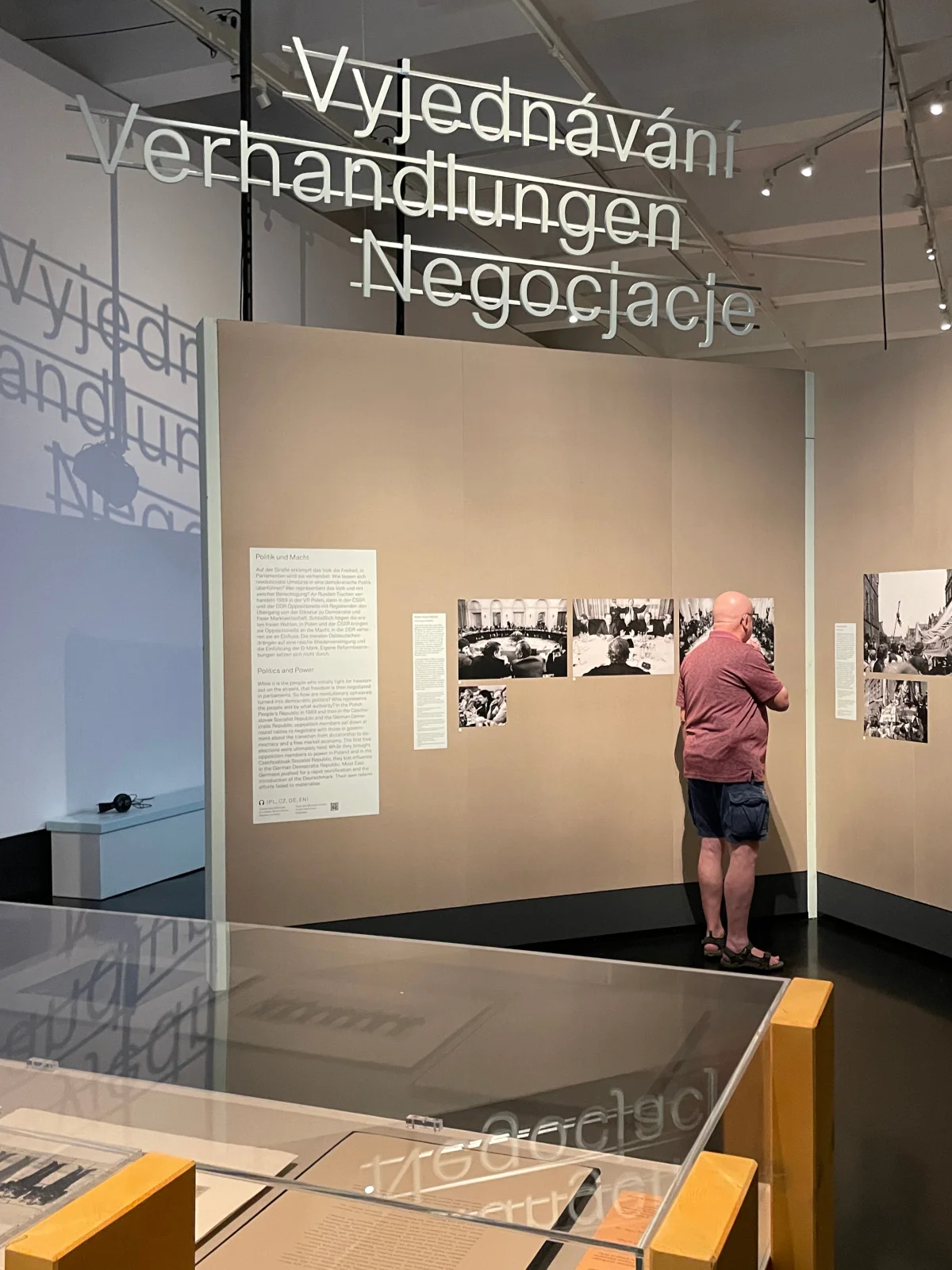 Eine Person betrachtet Fotografien an einer Ausstellungswand unter dem dreisprachigen hängenden Titel „Verhandlungen“.