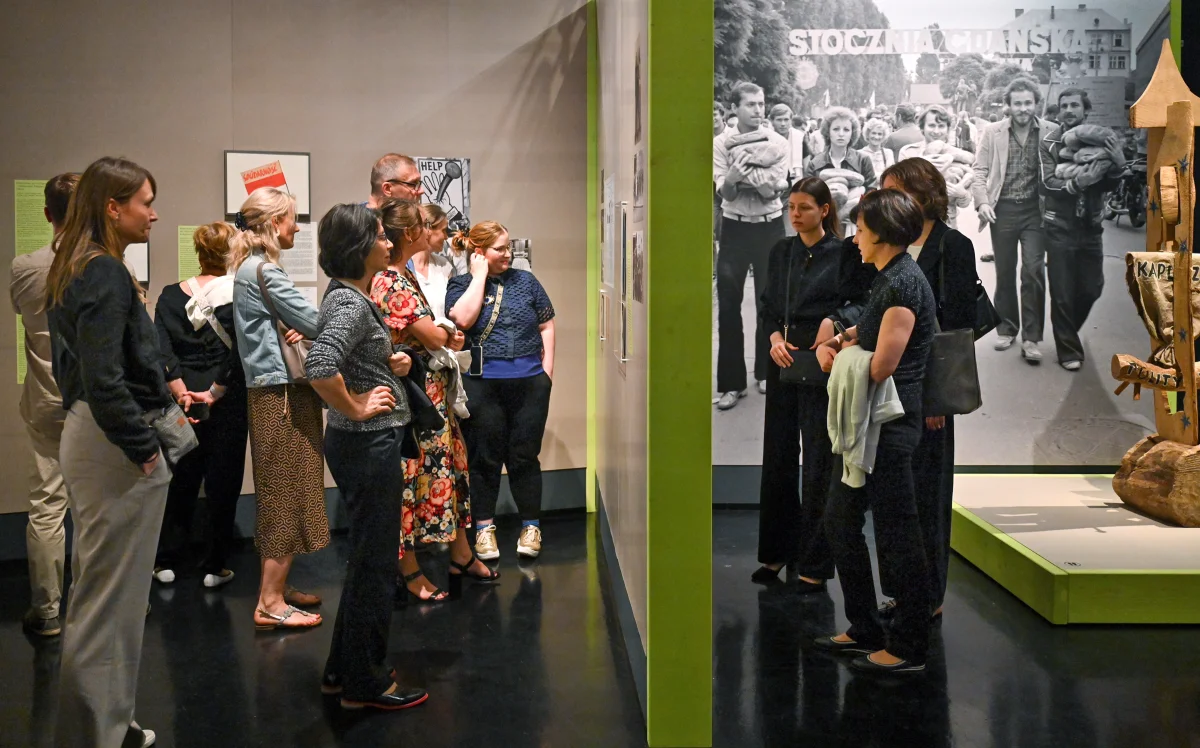 Eine Besuchergruppe steht zwischen einer grünen Ausstellungswand, einem historischen Gruppenfoto und einer geschnitzten Skulptur.