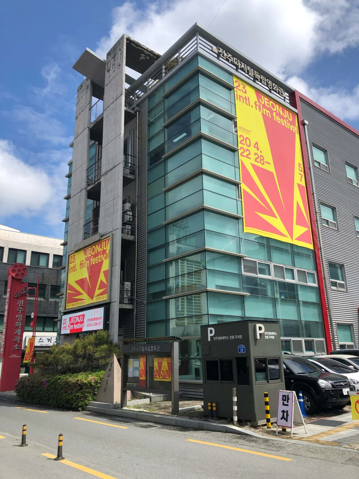 An der Ecke eines mehrgeschossigen Gebäudes mit blauer Glasfassade hängt ein großes rot-gelbes JEONJU-Festivalbanner.