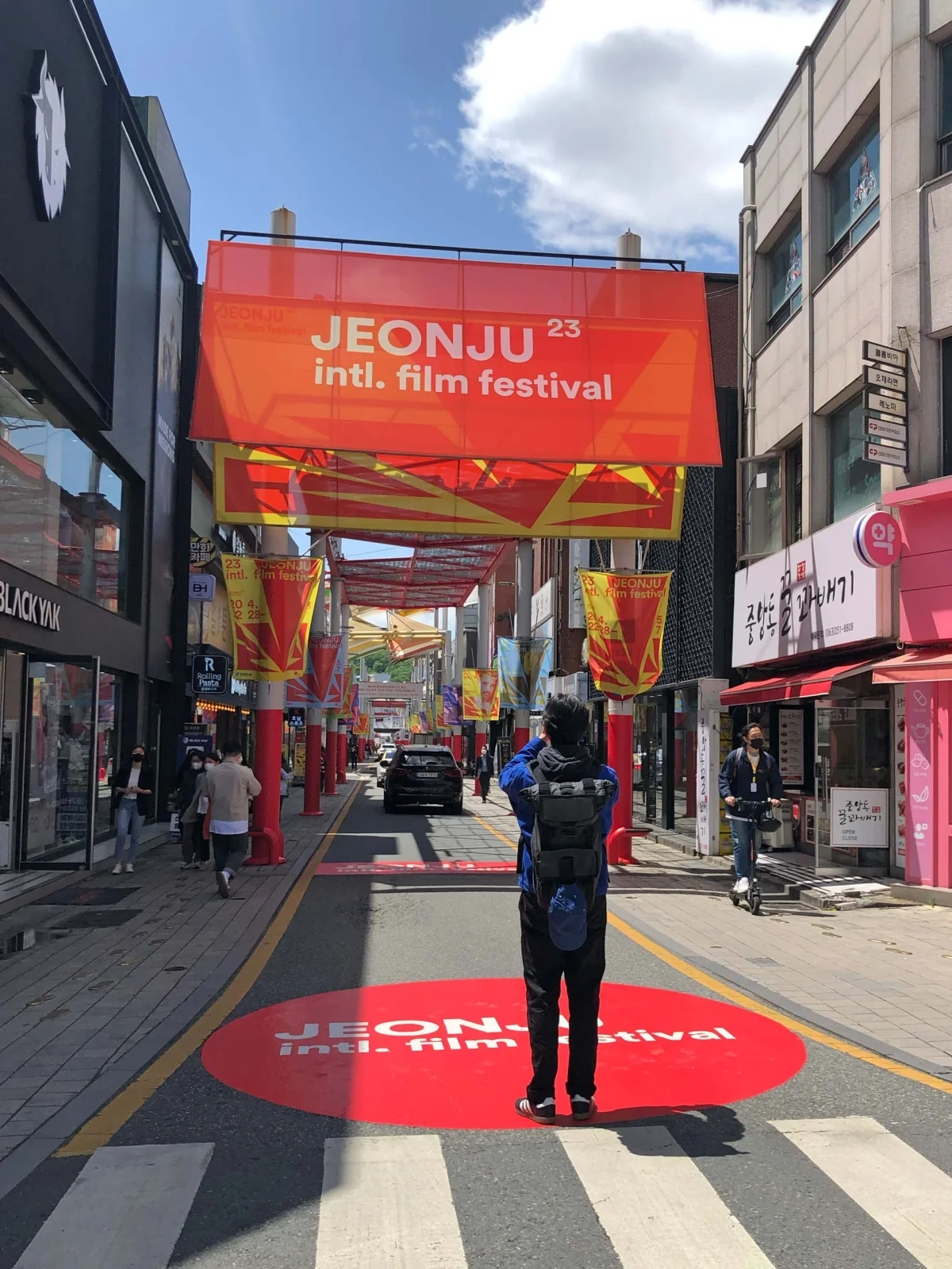 Über einem Straßeneingang hängen ein orangefarbenes JEONJU-Banner und eine rot-gelbe Stoffüberdachung; davor steht eine Person mit Kamera.