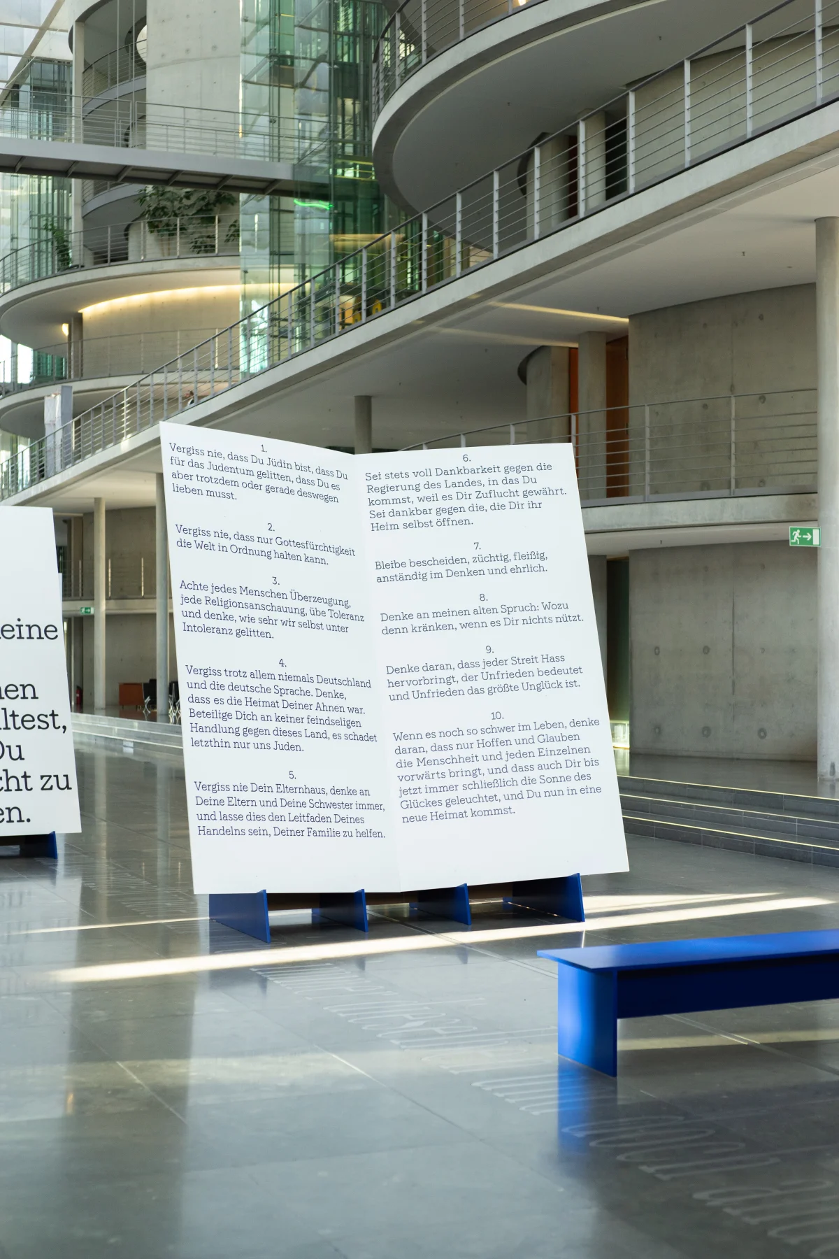 Freestanding white exhibition module with a bilingual introduction in an atrium, with a blue bench in front.
