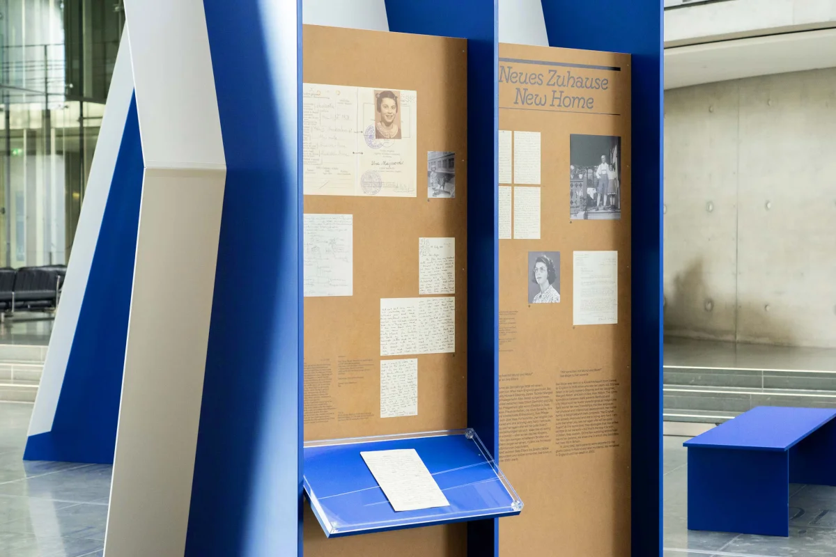 Blue exhibition modules with historical documents and photographs; one panel is headed “Neues Zuhause / New Home”.