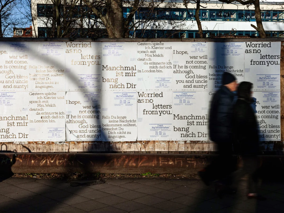 Wall covered with closely arranged exhibition posters and large letter quotations, with a person walking past.