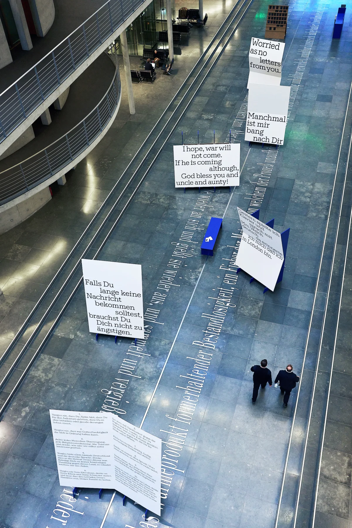 Overhead view of the Kindertransport exhibition, with white text panels, blue benches and visitors.