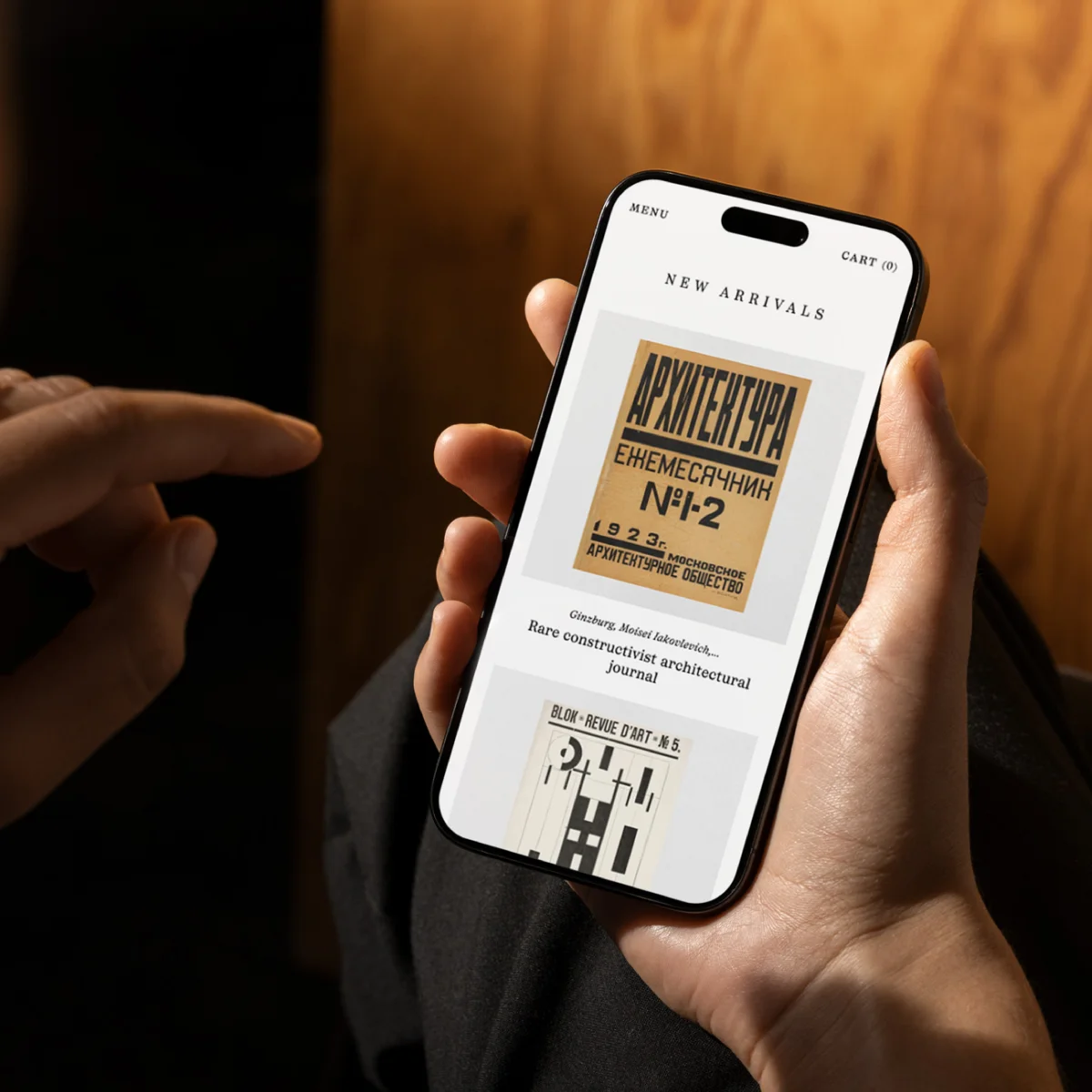 Hände halten ein Smartphone mit der PENKA-Seite „New Arrivals“, auf der historische Buch- und Zeitschriftencover stehen.