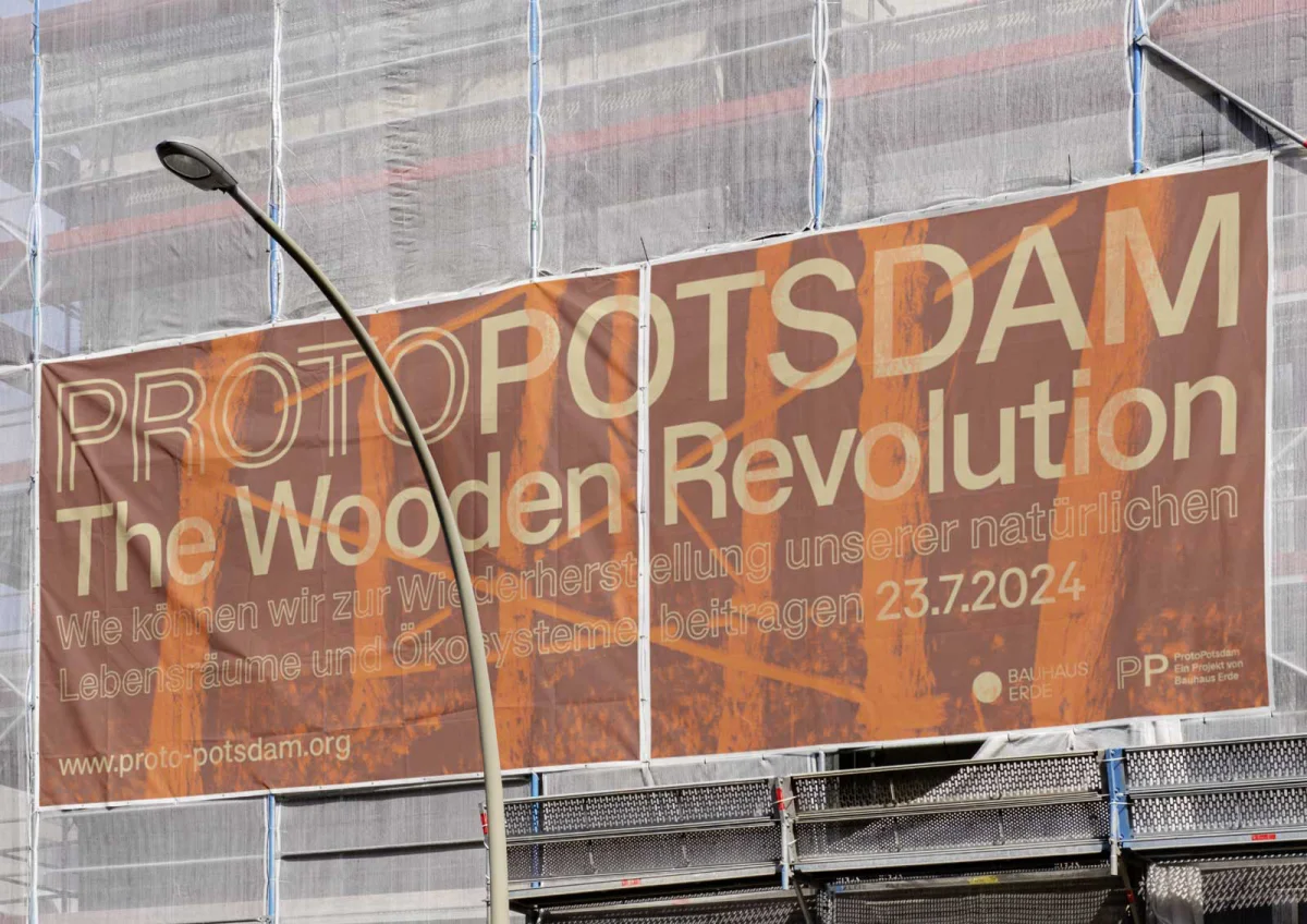 Large orange-brown construction banner reading “ProtoPotsdam – The Wooden Revolution” on a scaffolded facade.
