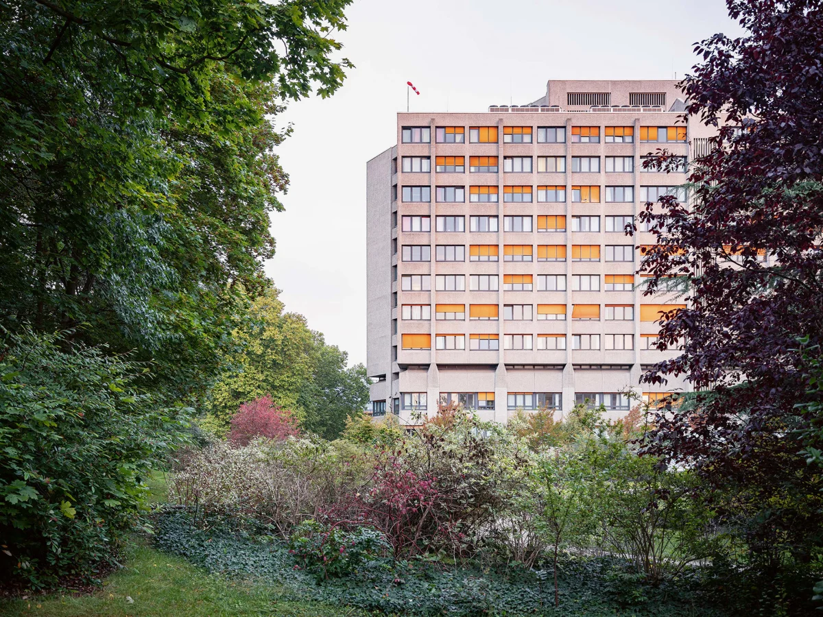 Contemporary view of a tall building with regular rows of windows and orange accents behind dense greenery.