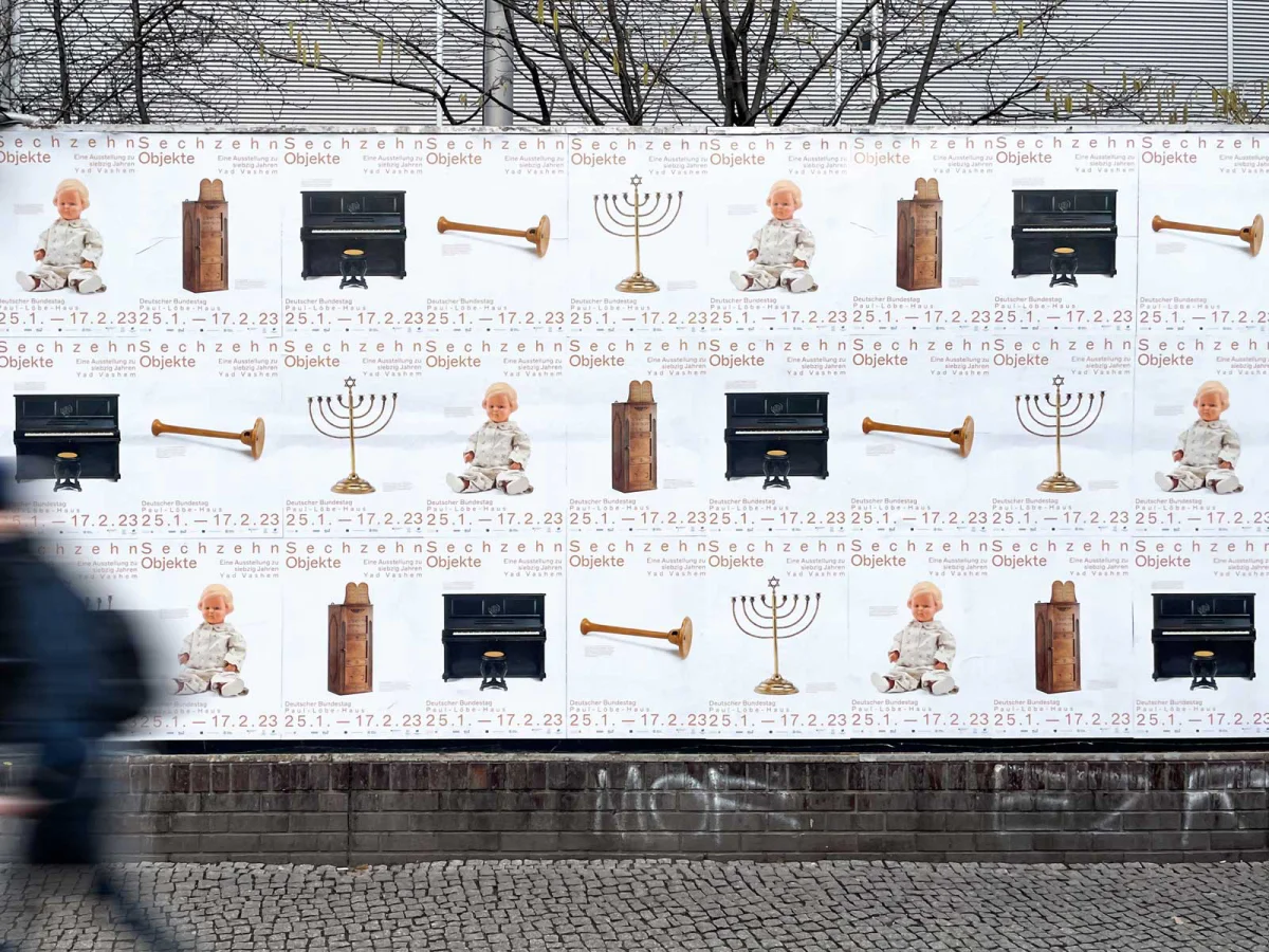 Street wall covered with repeated “Sixteen Objects” posters featuring a doll, Torah ark, piano, wind instrument and menorah.