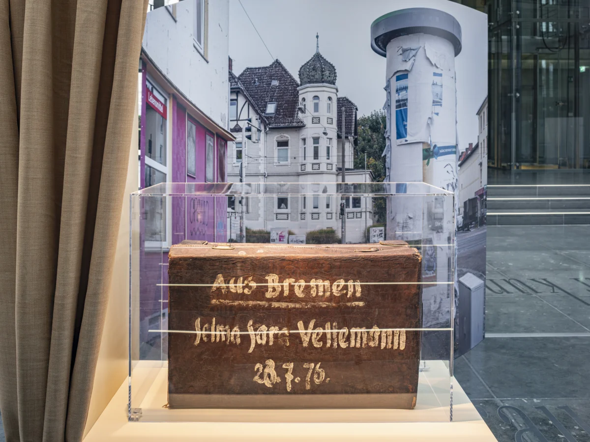 Inscribed historical wooden box in a transparent case in front of a large photograph of houses.