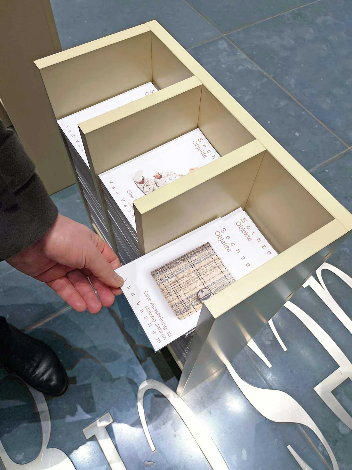 Hand taking a “Sixteen Objects” booklet from an exhibition stand divided into three compartments.
