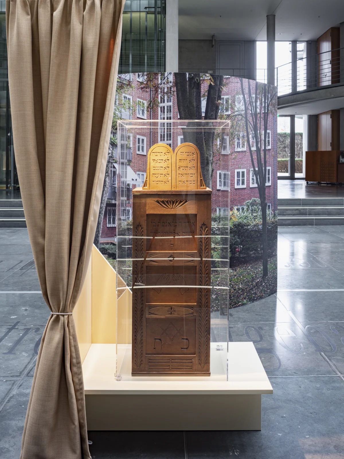 Inscribed wooden Torah ark in a display case, framed by a beige curtain and a building photograph.