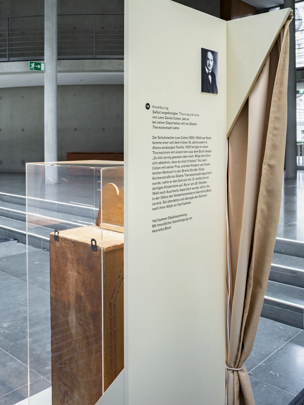 Side view of an exhibition module with a portrait and biographical text beside a display case containing a wooden Torah ark.