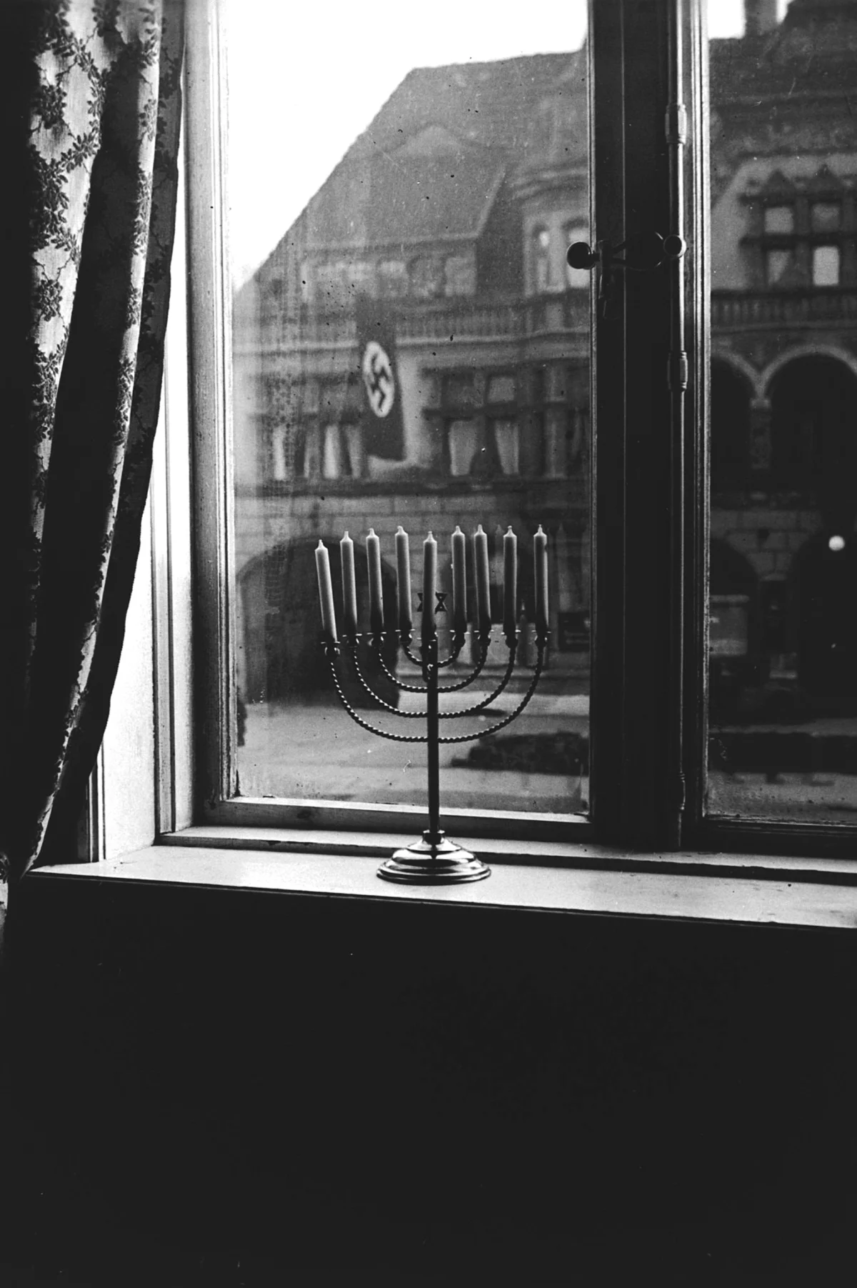 Historical black-and-white photograph of a Hanukkah menorah in a window, with a swastika flag on the building opposite.
