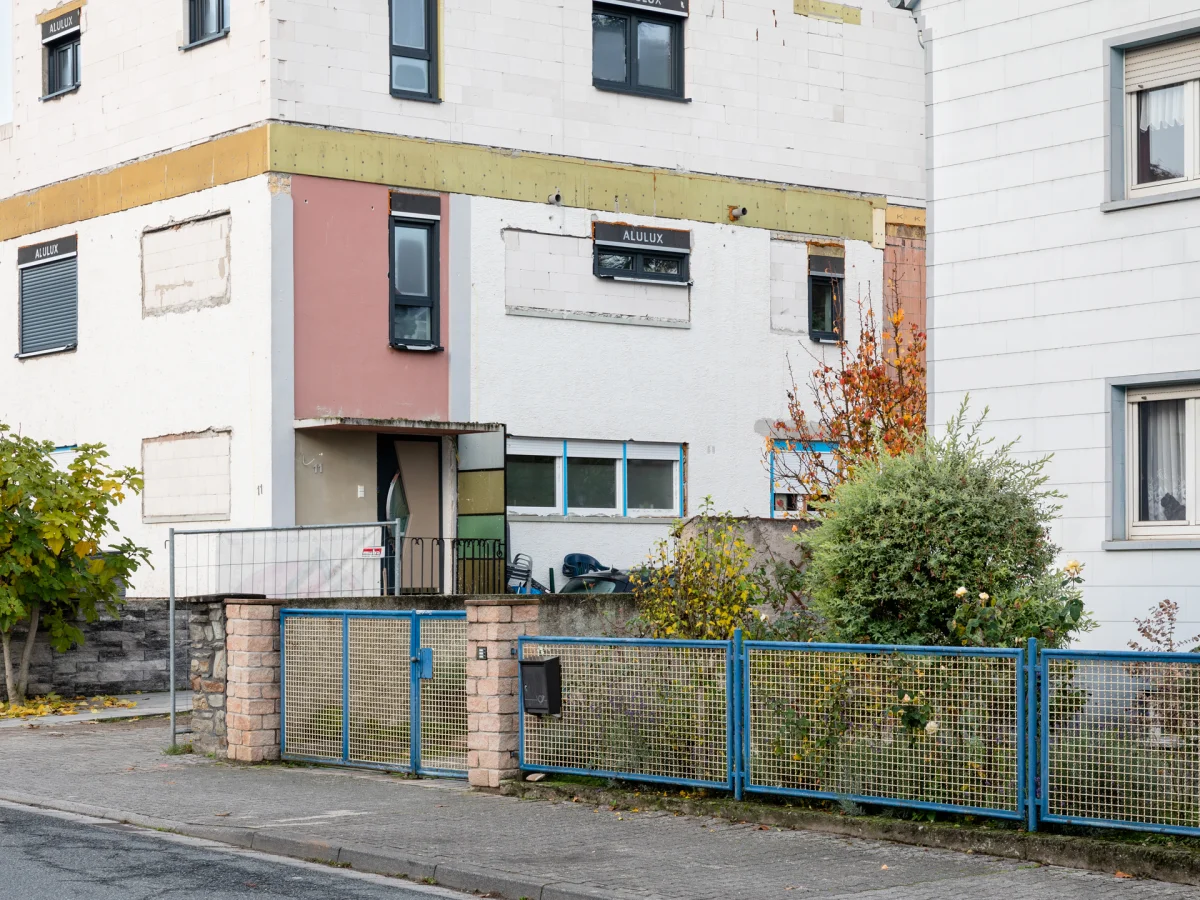 Residential building with a partly stripped facade, differently coloured wall areas and a blue-and-yellow garden fence.