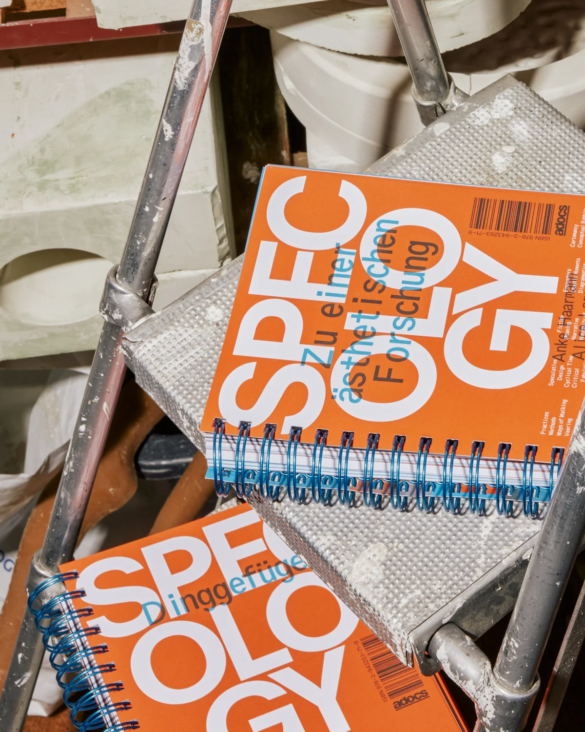 Two orange “Specology” volumes on a metal stepladder, with blue spiral binding connecting the chapters.