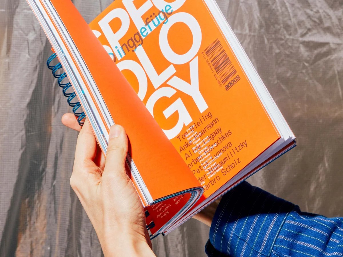Hands holding “Specology”, with an orange cover, large white typography and blue spiral binding.