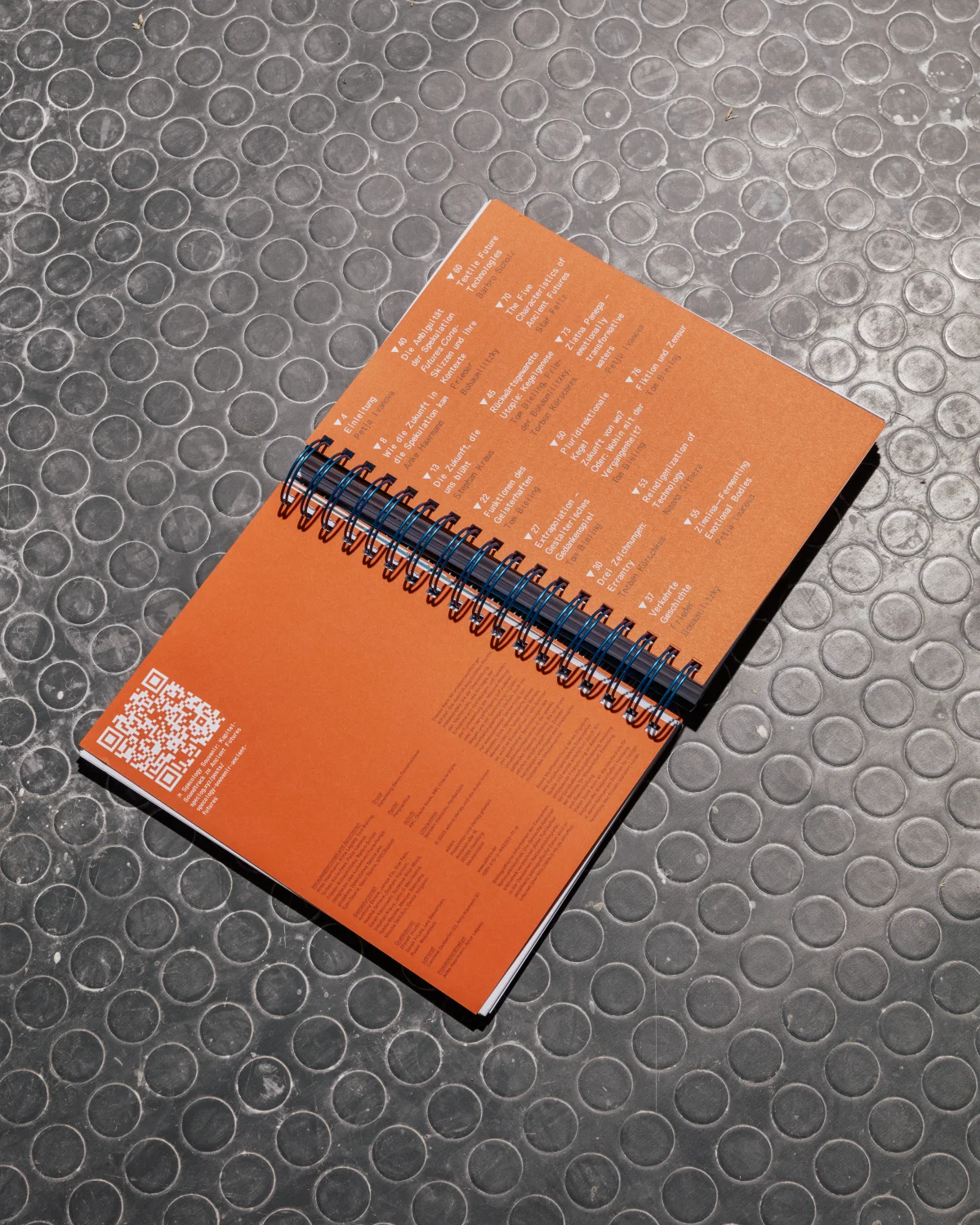 Opened orange back-cover panels of “Specology”, with index text, a QR code and central spiral binding.