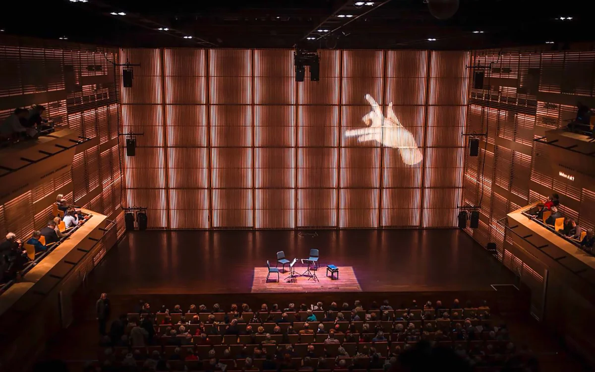 Concert hall with an audience and stage, with a hand image projected onto the large rear wall.