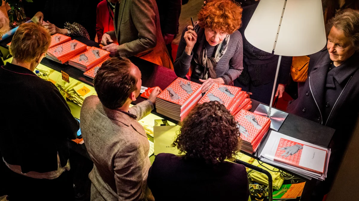 People at a sales table with stacks of red Biennale programmes and other printed materials.