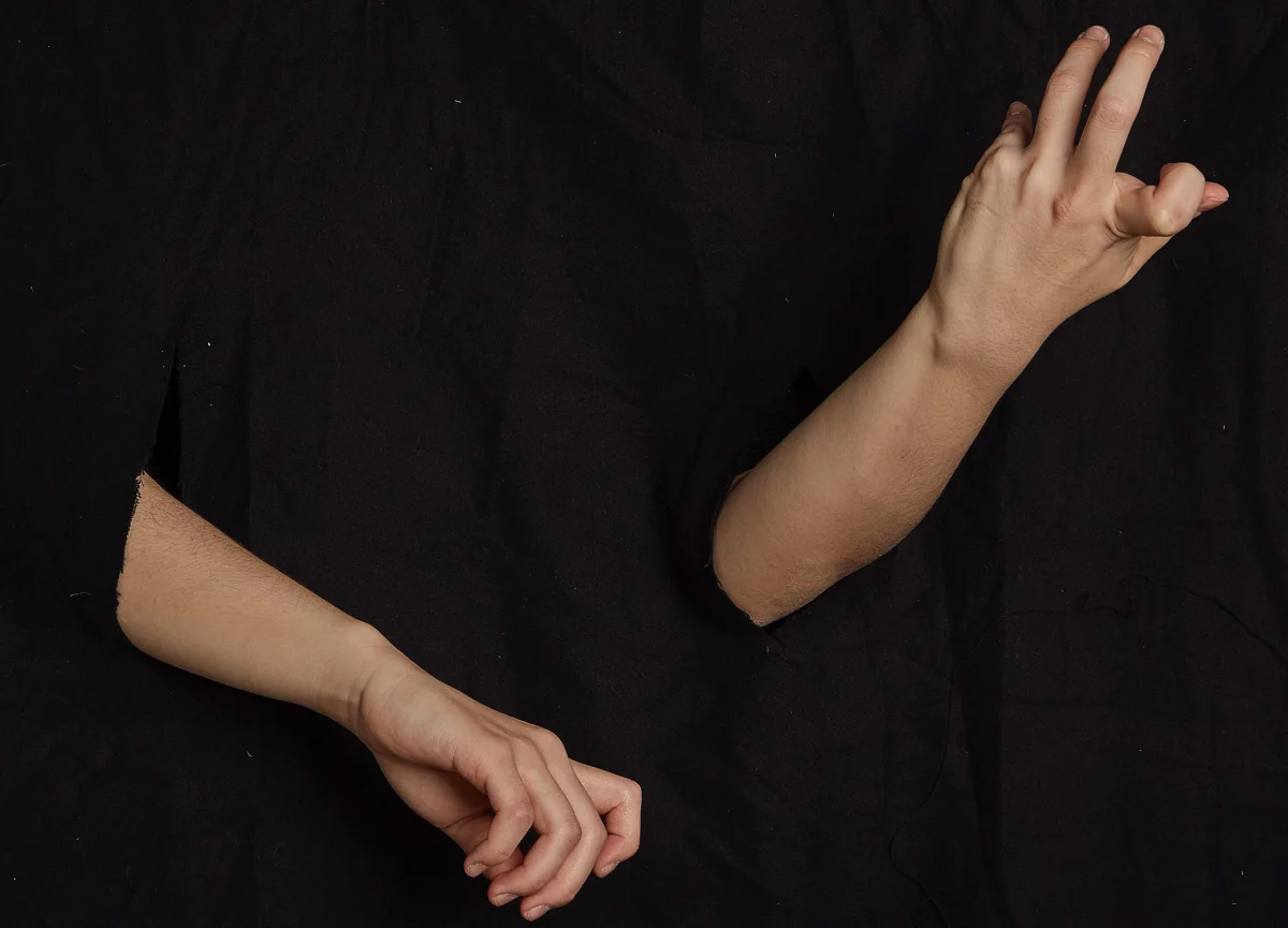 Arms and hands against black fabric, with one hand raised and the other pointing down with curved fingers.