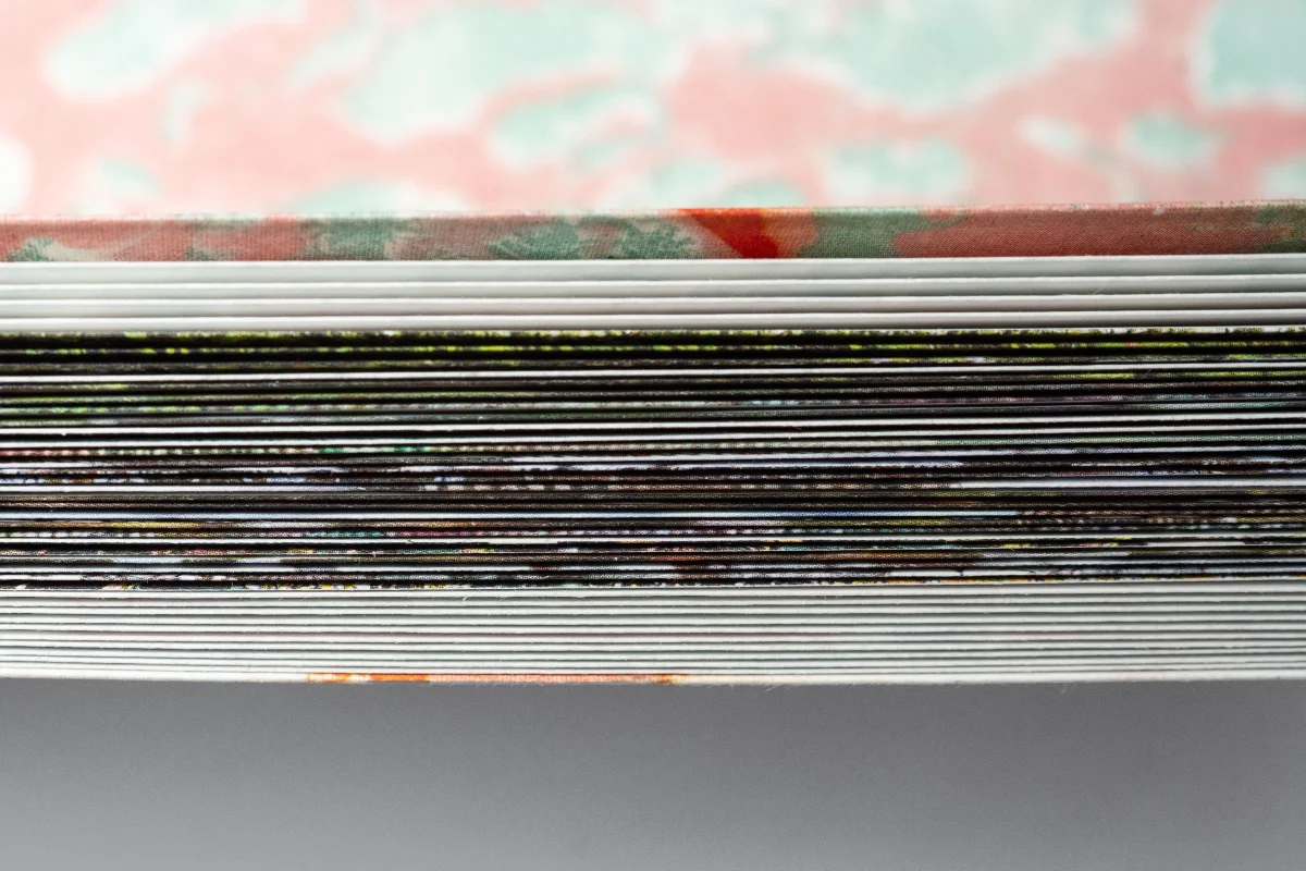 Side view of stacked, folded book pages with black photographic bands and a blurred pink and green cover.